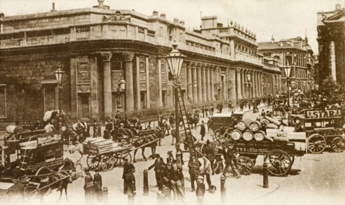 Outside the Bank of England. Horse-drawn carriages and pedestrians bustle about on a street running
