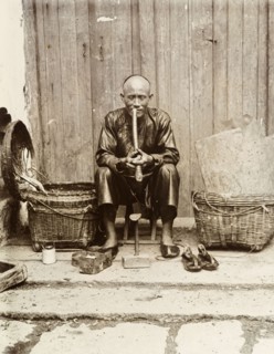 An itinerant shoemaker, Hong Kong. An itinerant shoemaker sits on a stool, smoking a long pipe at