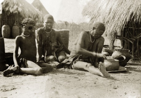 Preparing a meal. A woman and child sit on the ground outside a village hut, watching as an older