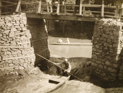 Shooting the rapids after the flood'. A man steers a canoe beneath a footbridge through rapids that
