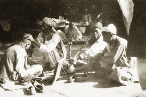 Barber's stall on a street in Peshawar. A man sits cross-legged as he has his head shaved at a