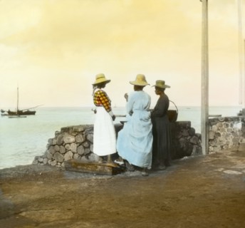 On the waterfront at Roseau. Three women in straw hats and dresses chat together on the waterfront