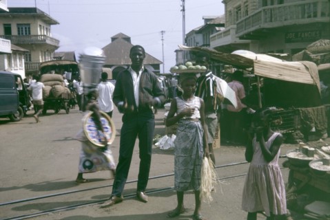 Market scene at Freetown, Sierra Leone. A woman balances a platter of fruit on her head, as she