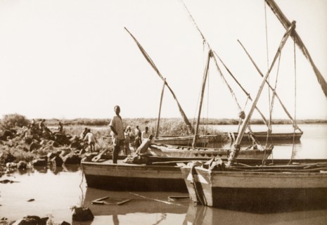 Sailboats moored in shallow water. A man stands at the stern of one of several small sailboats,