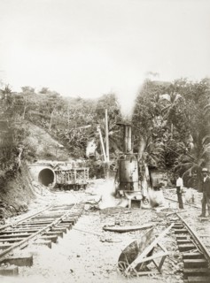 Railway construction site. Labourers stand on the tracks beside steam-operated machinery at a