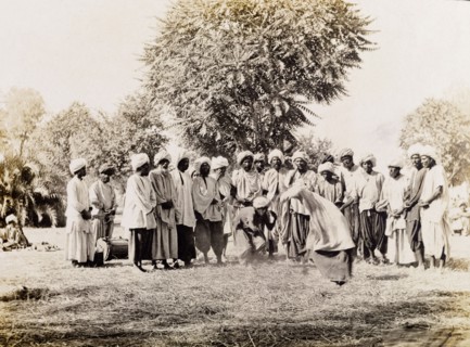 A wrestling match in Sukkur . Men in traditional Indian dress stand around a straw-covered patch of