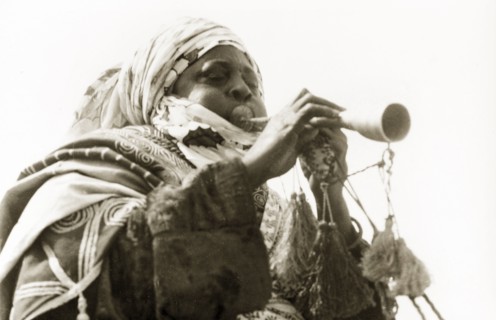 A Nigerian musician. A Nigerian musician plays a trumpet decorated with tassels. Nigeria, circa