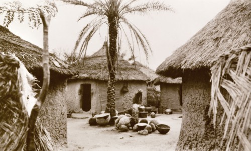 Interior of a Nigerian village. A woman undertakes domestic chores outdoors in the centre of a