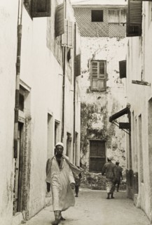 A back street of Lamu. A man in traditional Arab dress, including a kurta (shirt) and a fez, walks