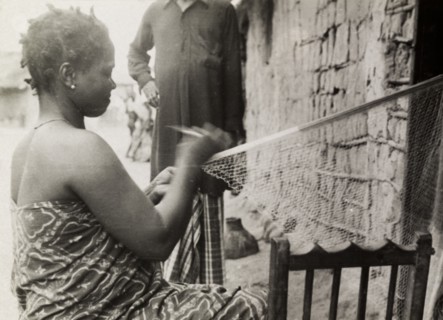 Repairing fishing nets. A Ghanaian woman in traditional dress sits outside a mud-walled building as