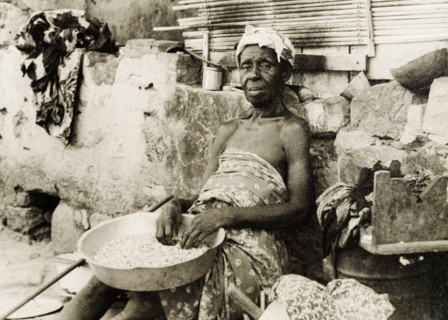 An elderly street vendor in Accra. An elderly woman sits against a roadside wall, selling nuts or