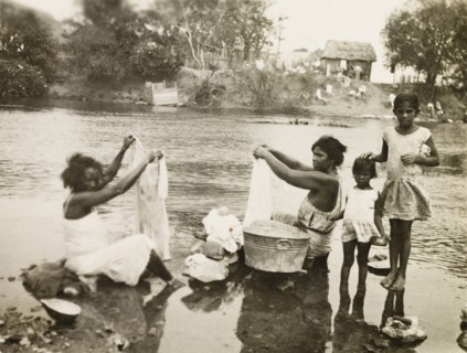 Washing clothes in a stream. Two female plantation workers, accompanied by two young girls, sit in