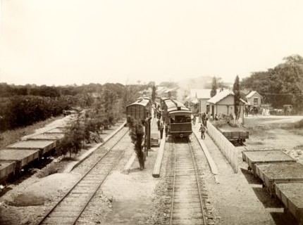 San Joseph railway station, Trinidad. Flatbed freight cars loaded with gravel sit beside passenger