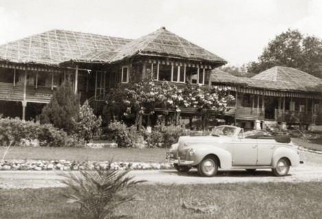 A stilted bungalow in British Malaya. A small car is parked in the driveway outside Dr Tweedie's