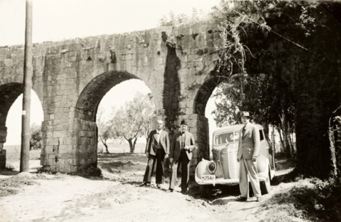 Visiting the aqueduct near Acre. Three men in suits stand beside a car beneath the arches of the
