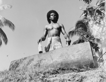A Fijian drummer with a 'lali'. A Fijian man wearing a 'sulu' (wraparound skirt) stands over a