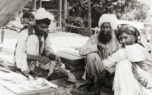 A visit to the cobbler. Two elderly men visit a cobbler who squats on the ground, mending boots in