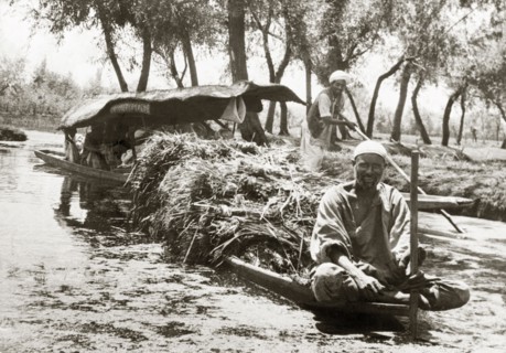 A canal dredger in Srinagar. A canal dredger sits cross-legged on the bow of a long canoe, which is