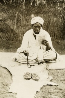Nigerian man praying. A Nigerian man sits cross-legged on a mat, holding prayer beads in one hand