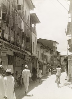 Commercial street in Stone Town. Tall, narrow shops with shuttered windows and awnings flank a