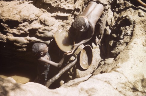 Waterhole in Mbaru. A young Suk (Pokot) woman stoops to collect a gourd full of water, passed to