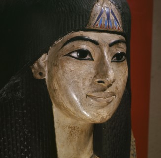 A mummy mask of a man wearing an elaborate collar and a heavy wig adorned with a lotus blossom