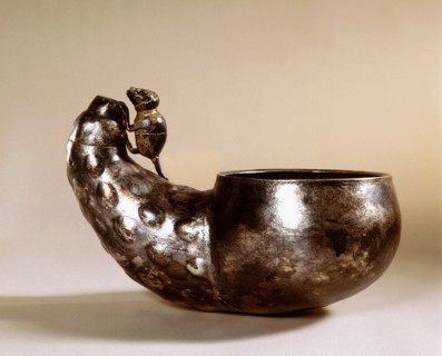 A cup made of beaten metal with a handle in the form of a mouse climbing over a vegetable