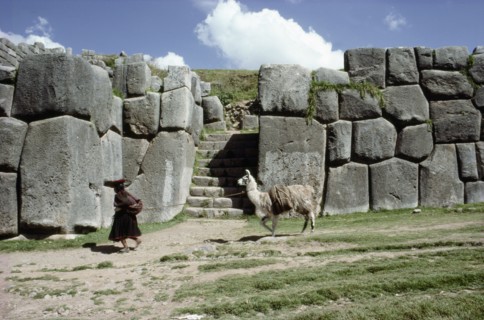 Quechua woman and llama walking past a monumental Inca doorway and wall at Sacsahuaman