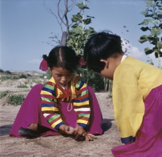 Two young Korean girls in brightly coloured national dress, crouching on the ground as they play a game with small stones