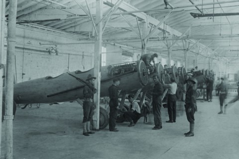 A line of airplanes is being assembled under the watchful eye of a gun-toting soldier as they are being completed to be sent to the European war; the factory is at College Point, Queens 1917