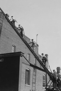A team of Firefighters with Hoses on Their backs climbs a ladder 1920