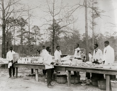 African American men preparing to serve a meal in an outdoor setting among trees 1900