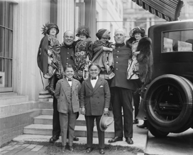Little People all dressed in their finery are lifted in the arms of full sized people. 1924