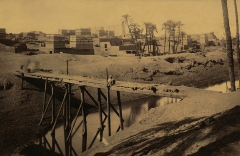 Man seated on wooden bridge over canal, ruins and buildings in background 1880