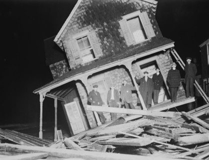 Men stand on Porch of a Sea bright Cottage torn from its foundation and tilted at a 45 degree angle.