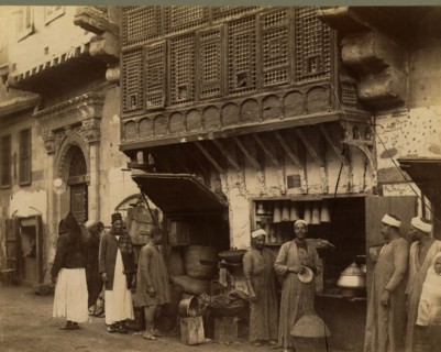 Men standing next to shop stall open to street, 1870
