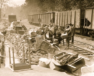 Men women and children flood victims pile up their home belongings to try to save them. They are all a train railroad signing with coal cars behind them and an embankment of a stream in front 1924