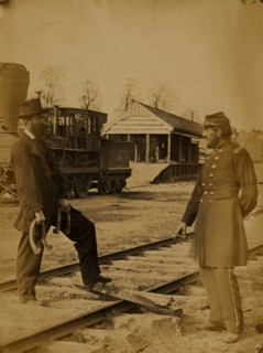 Soldier and another man standing at railroad tracks in front of locomotive 1863