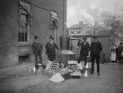 Onlookers watch as suited men stand in front of a large copper kettle still for making illegal liquor with boxes of bottles and funnels spread before them all for the manufacture of booze.
