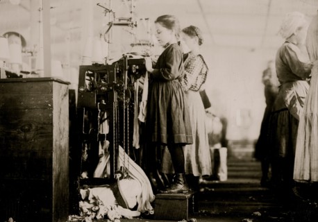 Knitter is a little girl so small she has to stand on a box to reach her machine. 1910