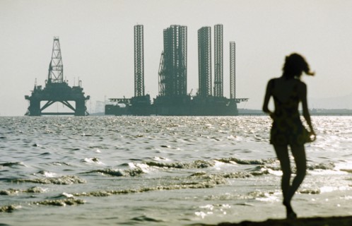 A woman in silhouette walking along shikhov beach on the shore of the caspian sea with an off-shore oil rig under construction in the background, baku, azerbaijan, october 2002.