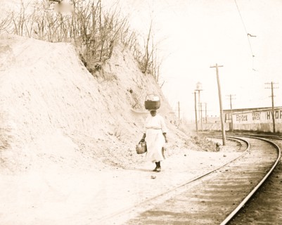 African American woman with basket on head walking near railroad tracks