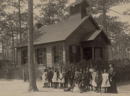 African American school children posed with their teacher outside a school, possibly in South Carolina