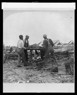 Galveston, Texas, 1900 - African-American men carrying a body on a stretcher, surrounded by wreckage of the hurricane and flood. Stereograph.