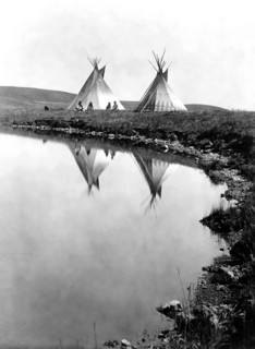 Edward S. Curits Native American Indians -  Two tepees reflected in water of pond, with four Piegan Indians seated circa 1910.