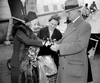 Senator William Gibbs McAdoo, of California, comes to the aid of Miss Marion Weldon, Paramount starlet, as she searches for her beauty aids before greeting the throng on her arrival at Washington Airp...