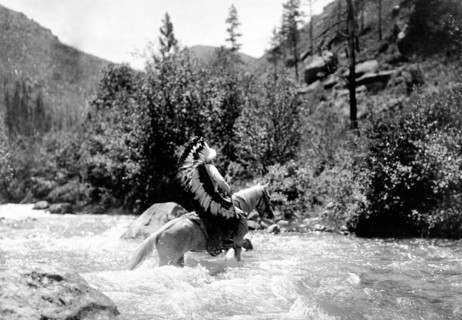 Edward S. Curtis Native American Indians - Bullchief, wearing warbonnet, crossing shallow rapids on horseback circa 1905.