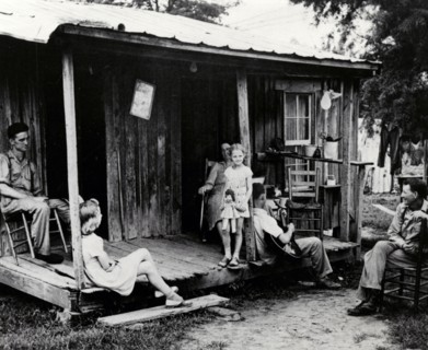 A rural family sits on an unscreened porch in the early evening