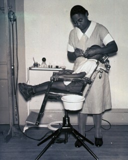 African American dentist is working on a patient's teeth in a sparsely furnished office.