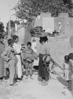 A man, street vendor, selling cakes in a Beirut street  to a crowd of young boys, man has cakes on top of his head ca. 1900